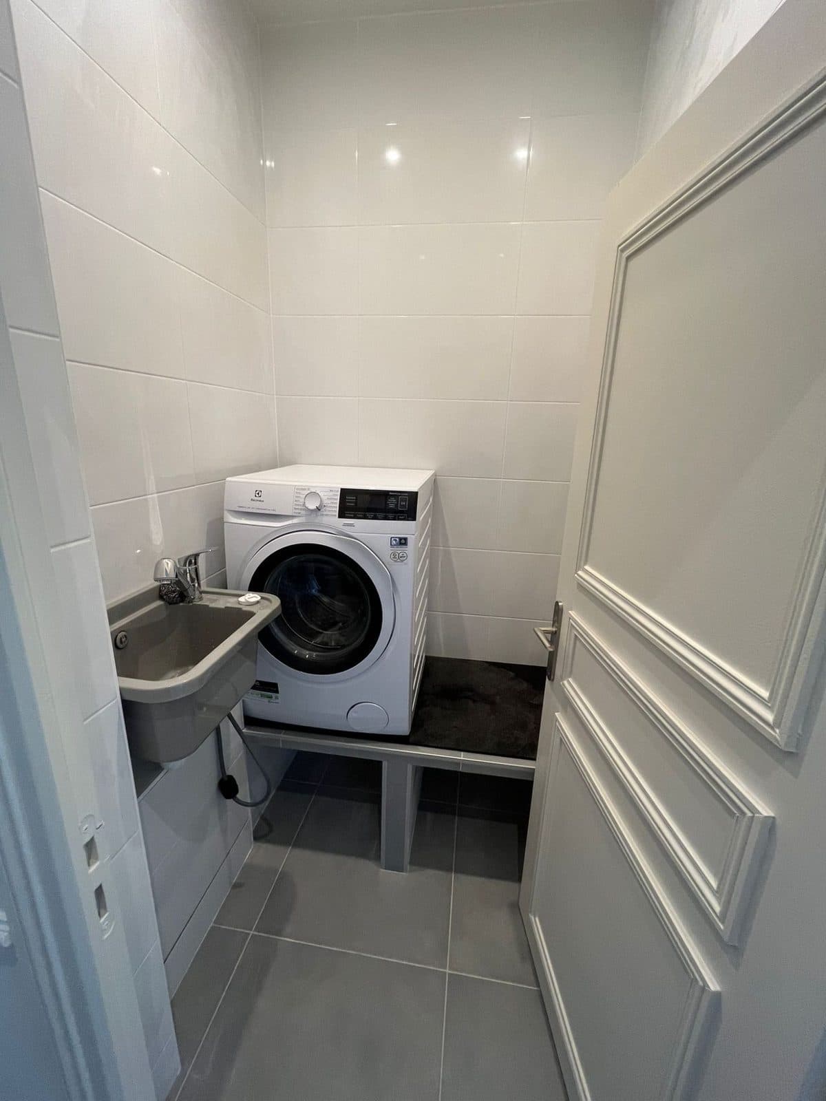 Buanderie : faïence blanche, sol gris, lave-linge surélevé sur un socle carrelé et bac à laver