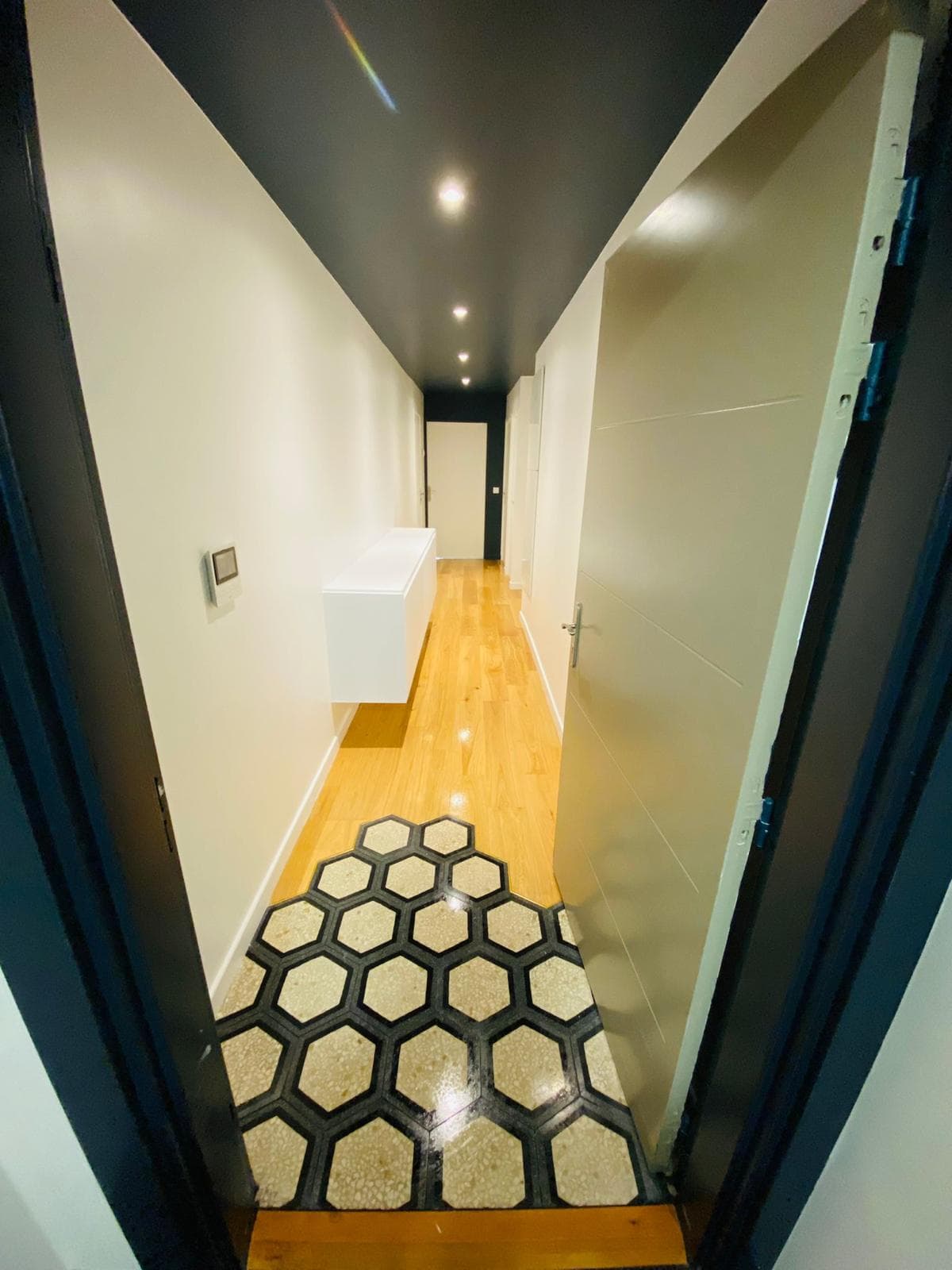 Couloir d'entrée : carreaux hexagonaux noir et beige qui se fondent dans un parquet en chêne, plafond noir avec spots encastrés