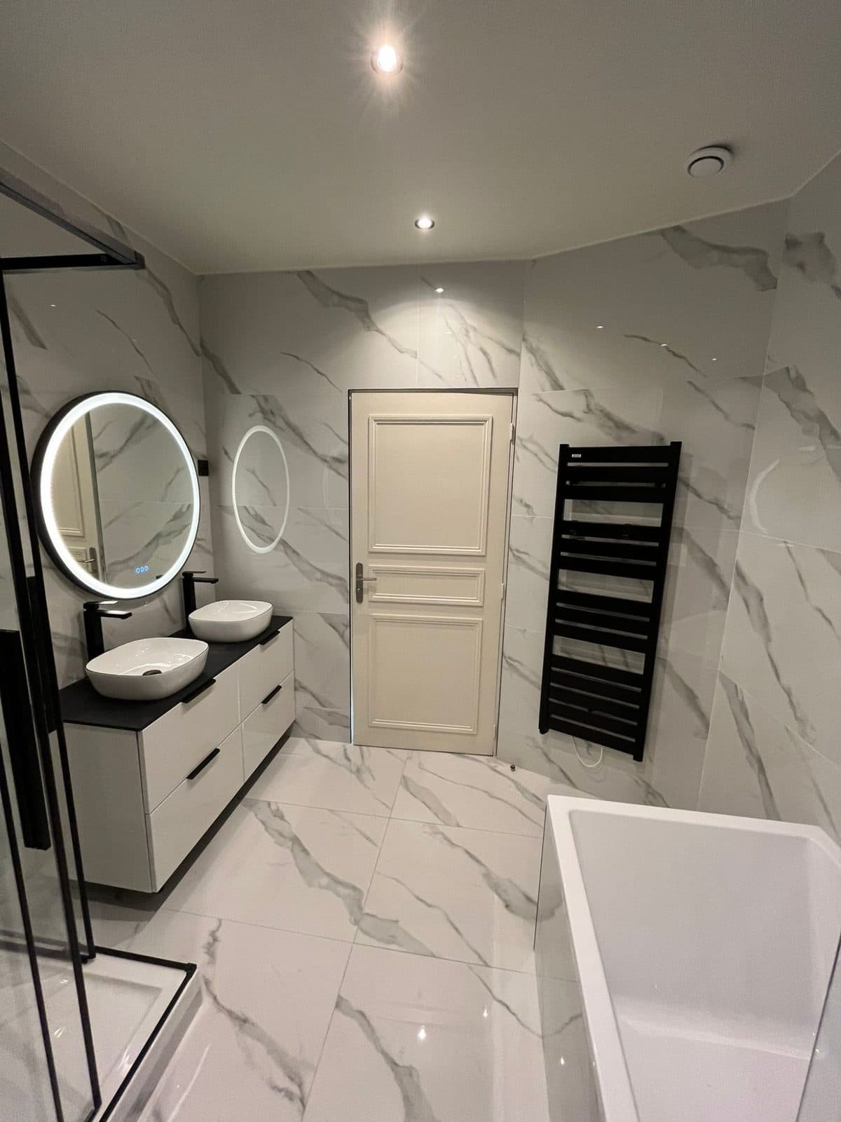 Salle de bain en grès effet marbre blanc : double vasque, meuble blanc et plan noir, miroirs ronds lumineux, sèche-serviettes noir