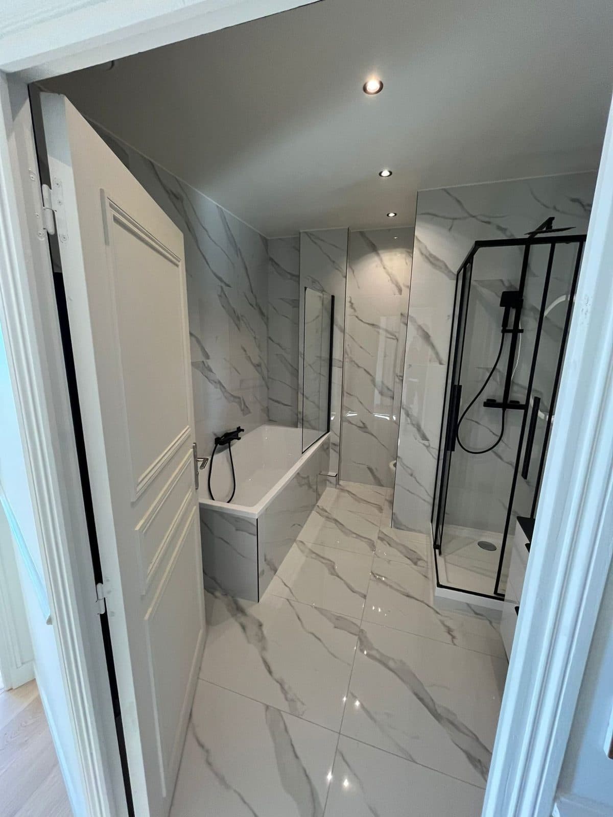 Salle de bain : baignoire avec pare-bain, douche à profilés noirs, murs et sol en grès effet marbre blanc