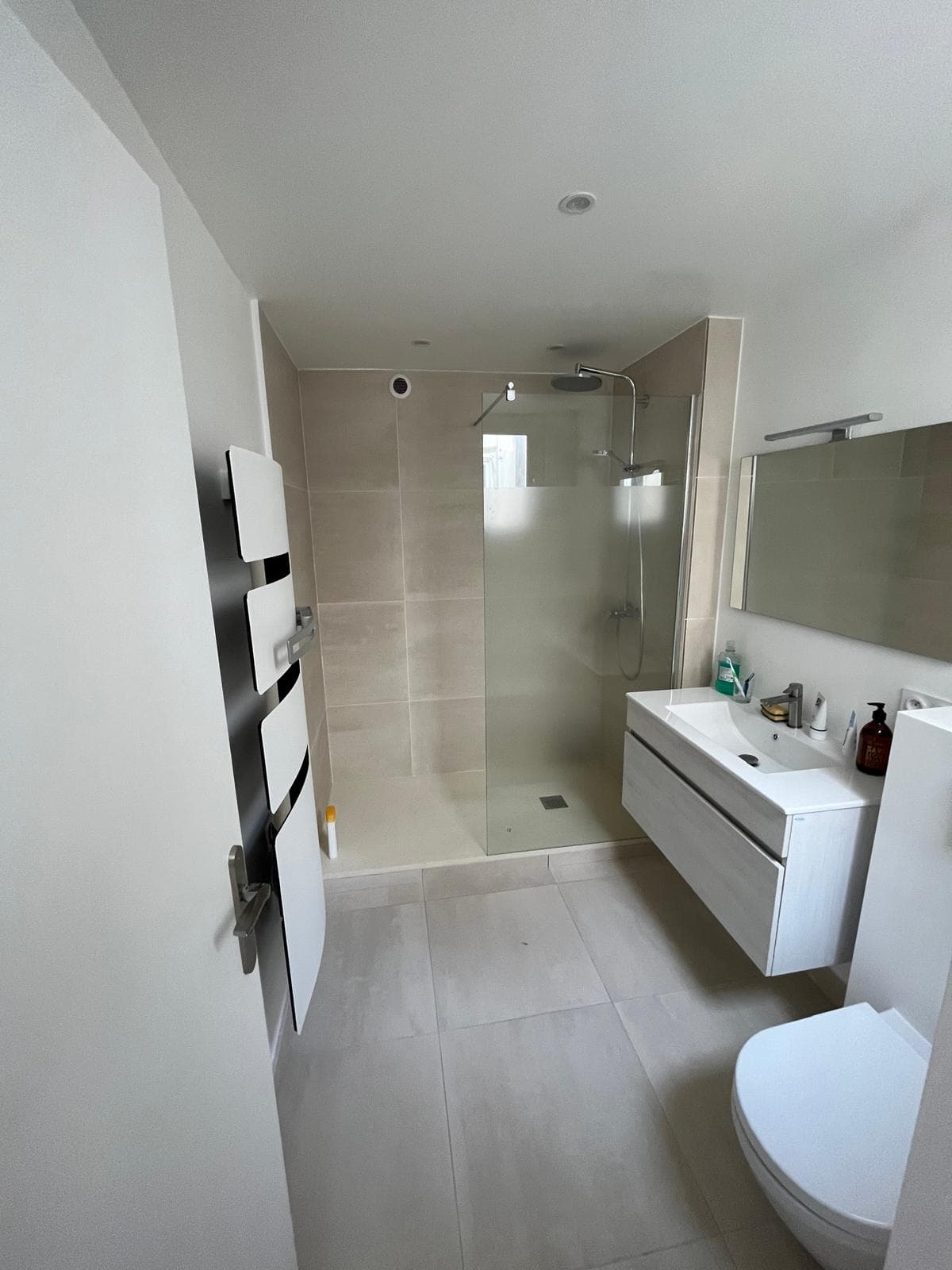 Salle d'eau rénovée : douche à l'italienne avec paroi vitrée, grands carreaux beiges, meuble vasque suspendu