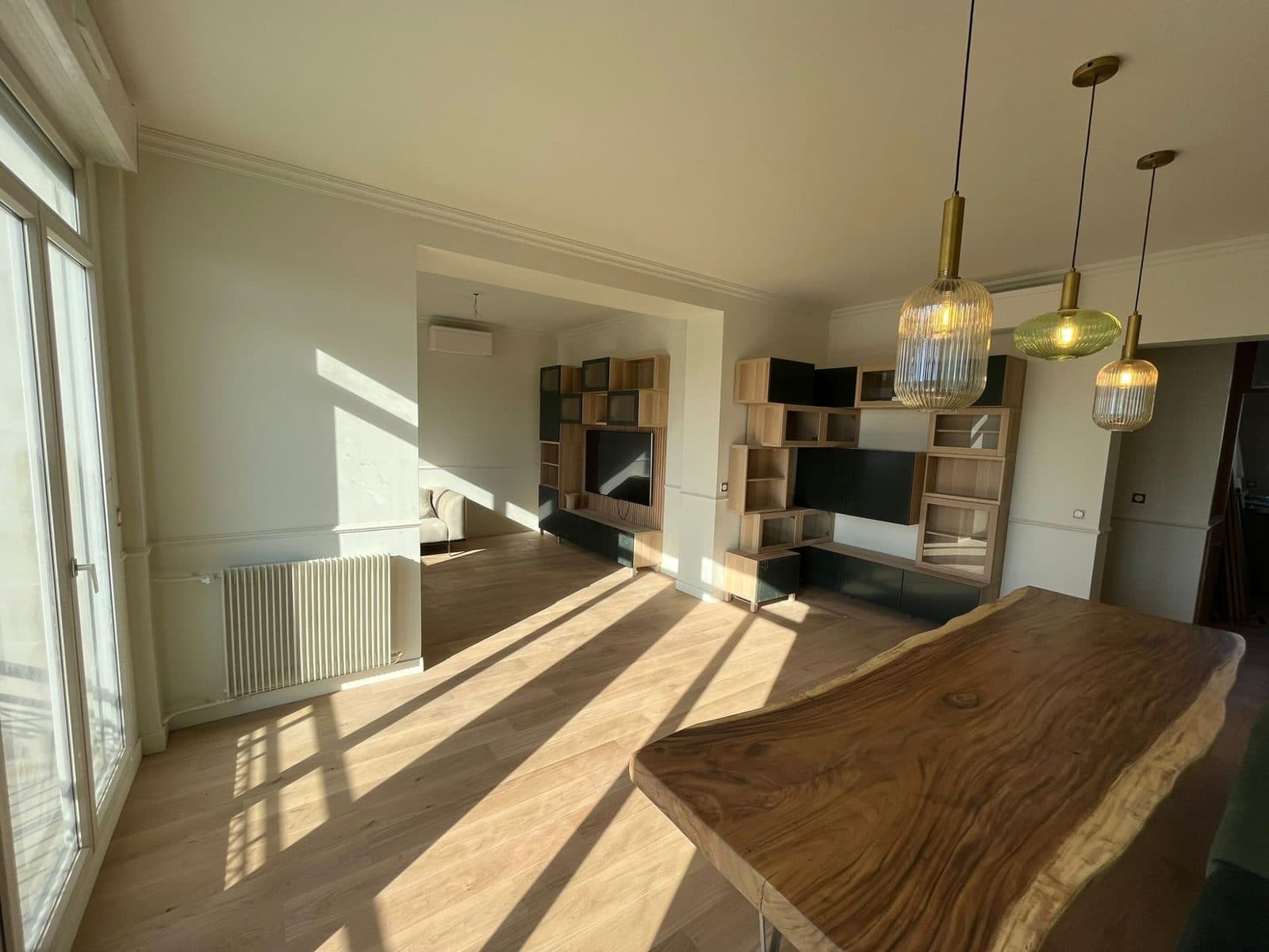 Séjour baigné de soleil : table en bois massif, trois suspensions en verre et laiton, bibliothèques en chêne et parquet clair